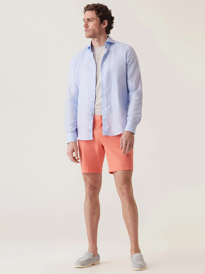 A man stands against a neutral background wearing a light blue button-up shirt over a white t-shirt, gray slip-on shoes, and the DEKE Galaxy Bermuda Ultra 4X Stretch Twill shorts in Sunset with a tailored fit.