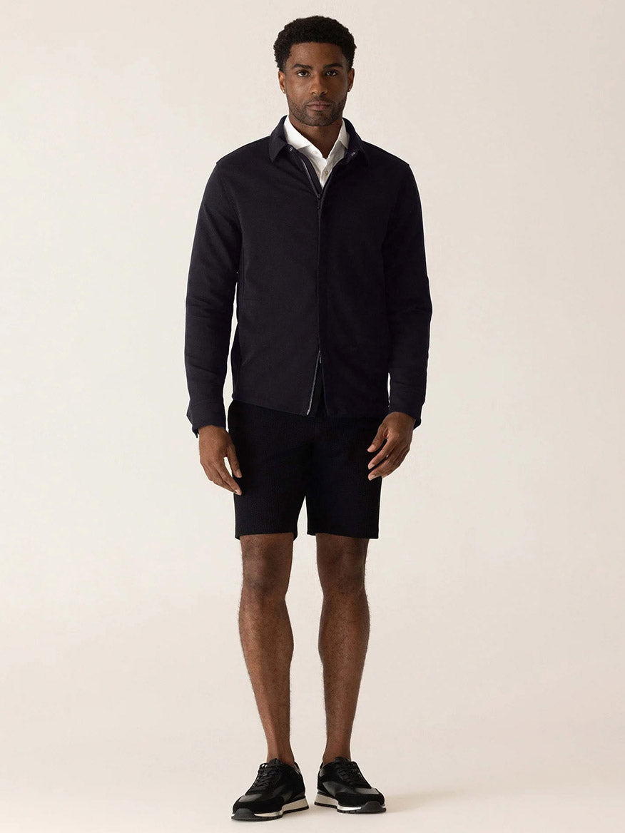 A man wears a black DEKE Getaway Shirt Jacket in Black Twilight Twill Jersey™ over a white button-down shirt, paired with black shorts and black sneakers.