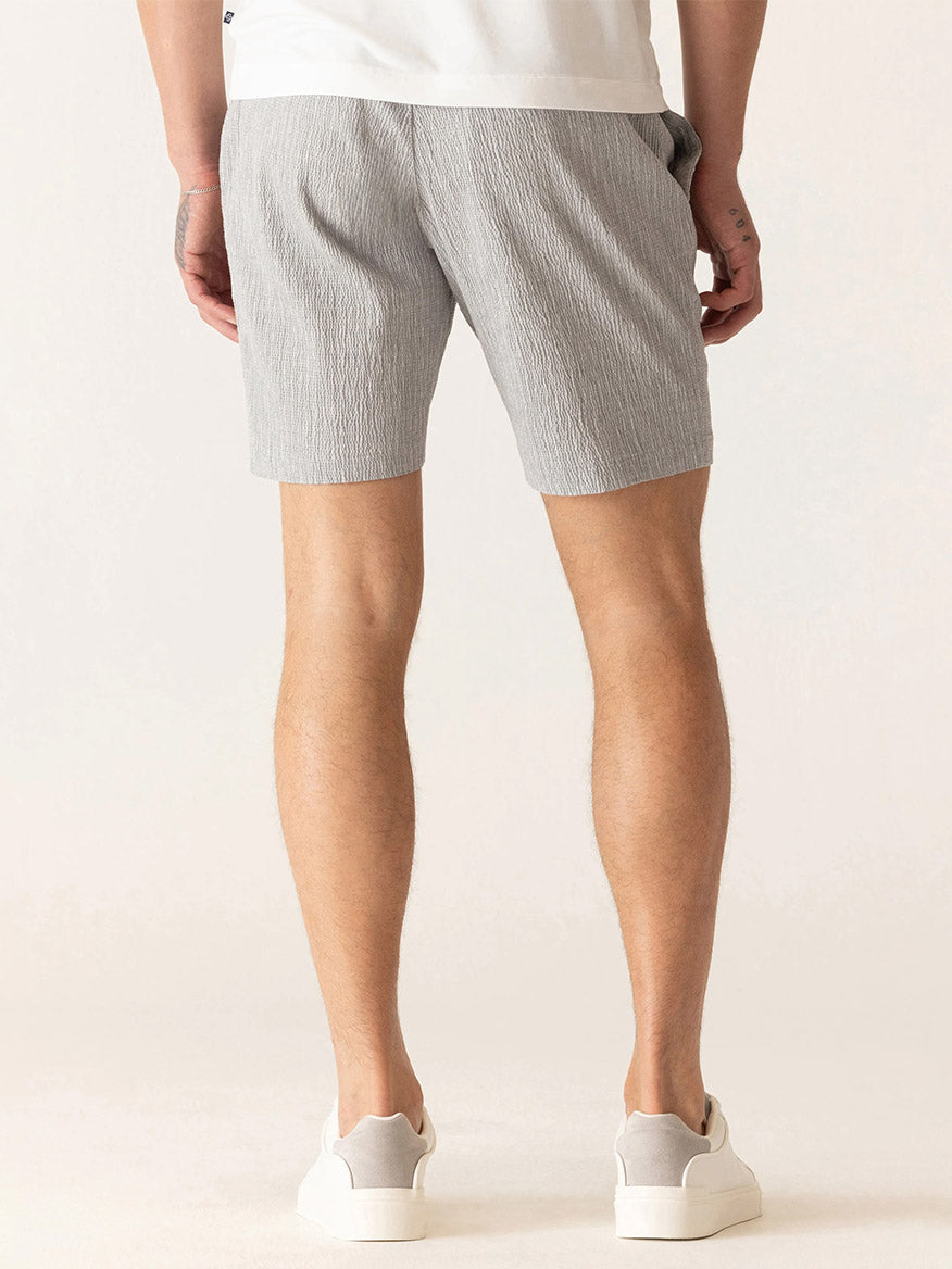 A person stands with their back to the camera, wearing a white t-shirt, DEKE Oasis Bermuda Supernatural Seersucker shorts in Grey/Natural, and white sneakers.