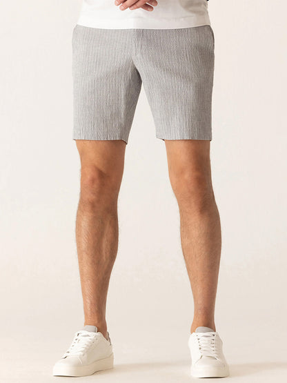 A person wears the DEKE Oasis Bermuda Supernatural Seersucker shorts in Grey/Natural, paired with a white shirt and white sneakers, standing against a plain background.