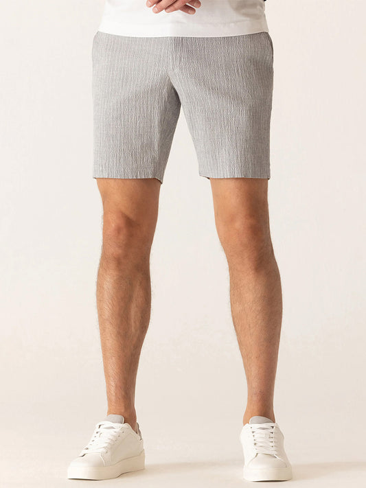A person wears the DEKE Oasis Bermuda Supernatural Seersucker shorts in Grey/Natural, paired with a white shirt and white sneakers, standing against a plain background.