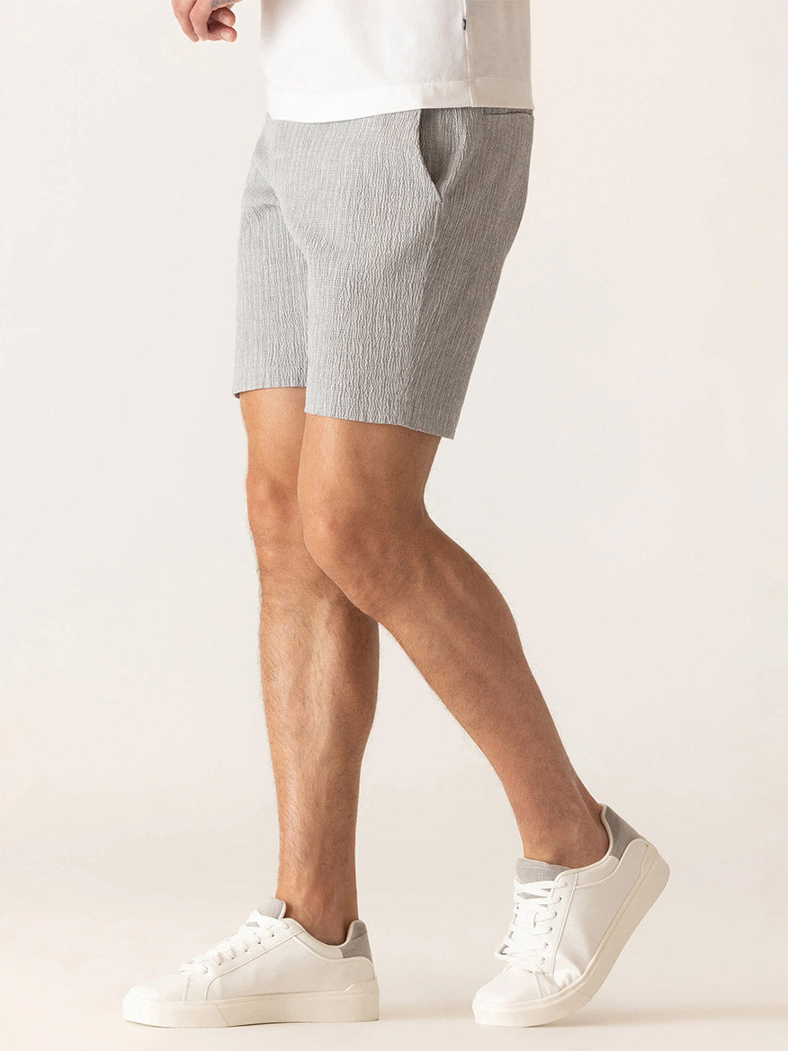 Man wearing DEKE Oasis Bermuda Supernatural Seersucker shorts in Grey/Natural, paired with a white t-shirt and white sneakers, standing against a plain light background.