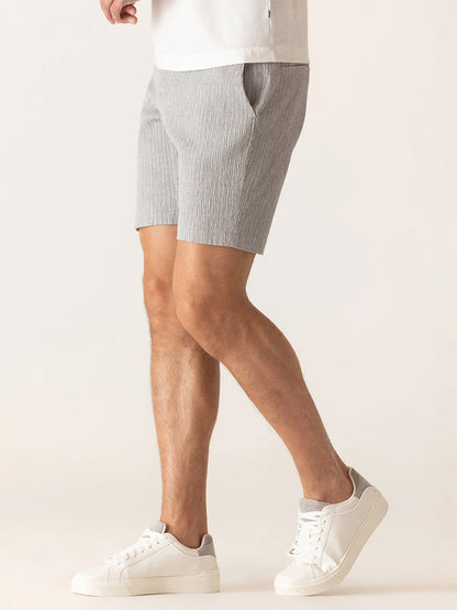 Man wearing DEKE Oasis Bermuda Supernatural Seersucker shorts in Grey/Natural, paired with a white t-shirt and white sneakers, standing against a plain light background.