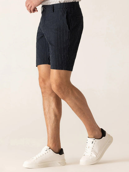 A person wearing DEKE Oasis Bermuda Supernatural Seersucker shorts in Midnight/Natural, a white shirt, and white sneakers stands against a plain light background.