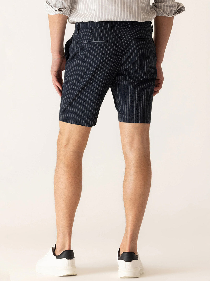 A person wearing DEKE Oasis Bermuda Supernatural Seersucker shorts in Midnight/Natural, a striped shirt, and white sneakers stands with their back to the camera.