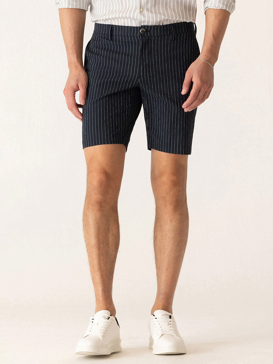 A man wears DEKE Oasis Bermuda Supernatural Seersucker shorts in Midnight/Natural, a light striped shirt, and white sneakers while standing against a plain light background.