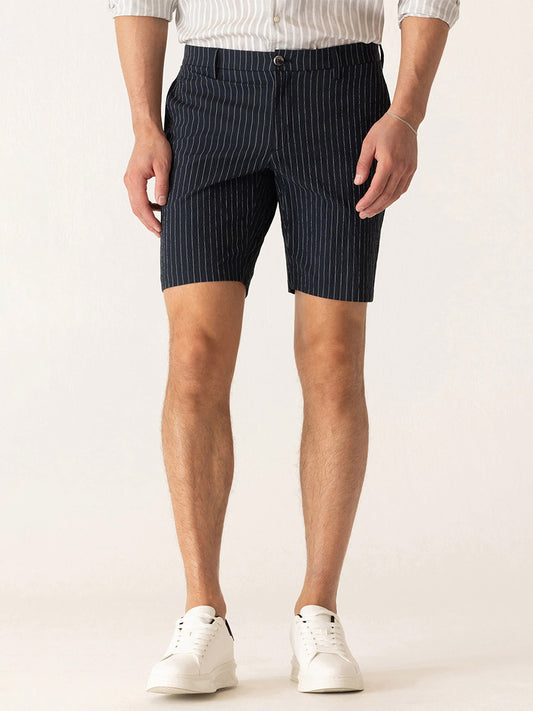 A man wears DEKE Oasis Bermuda Supernatural Seersucker shorts in Midnight/Natural, a light striped shirt, and white sneakers while standing against a plain light background.