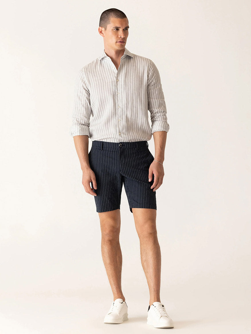 A man stands against a plain light background wearing a light striped long-sleeve shirt, white sneakers, and DEKE Oasis Bermuda Supernatural Seersucker shorts in Midnight/Natural.
