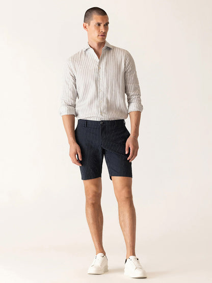 A man stands against a plain light background wearing a light striped long-sleeve shirt, white sneakers, and DEKE Oasis Bermuda Supernatural Seersucker shorts in Midnight/Natural.
