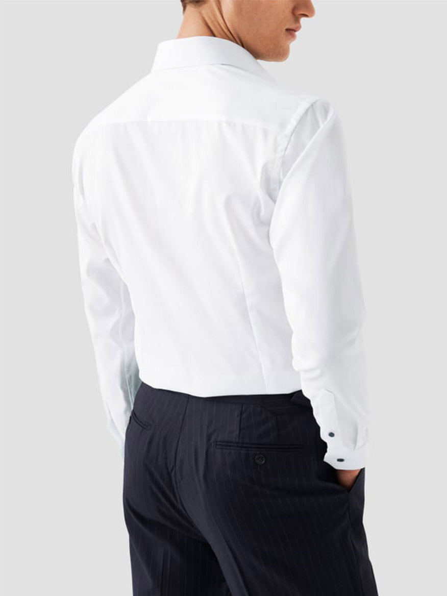 A man stands with his back to the camera against a plain light gray background, wearing the Eton Classic Fit Solid Signature Twill Dress Shirt in White with Navy Details and dark trousers.
