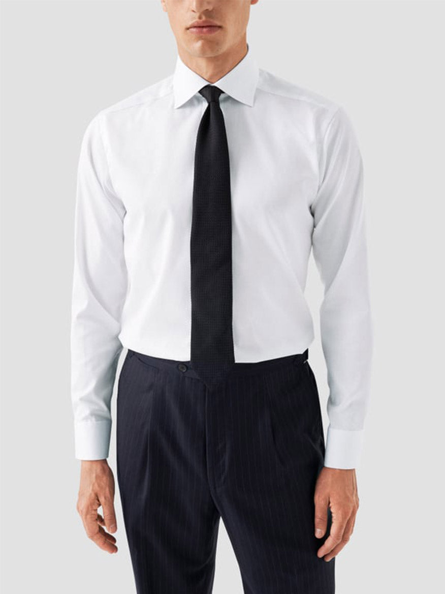 A man wearing an Eton Classic Fit Solid Signature Twill Dress Shirt in white with navy details, paired with a black tie and dark pinstripe trousers, stands against a plain light gray background.