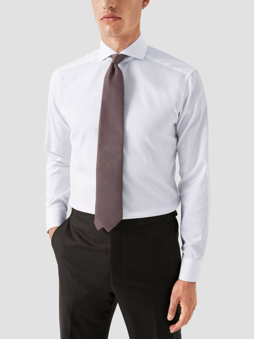 A man wearing an Eton Slim Fit Extreme Cut Away Dress Shirt in white, paired with a patterned brown tie and black trousers, stands with one hand in his pocket against a plain light gray background.