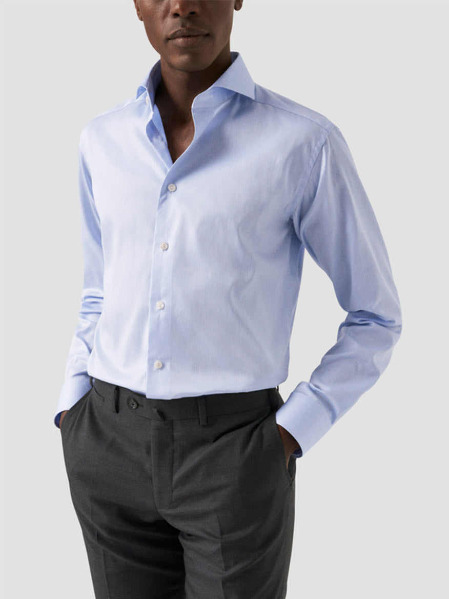 A person wears an Eton Slim Fit Extreme Cut Away Dress Shirt in Sky Blue, top button undone, paired with dark grey dress pants, hands in pockets, standing against a plain background.