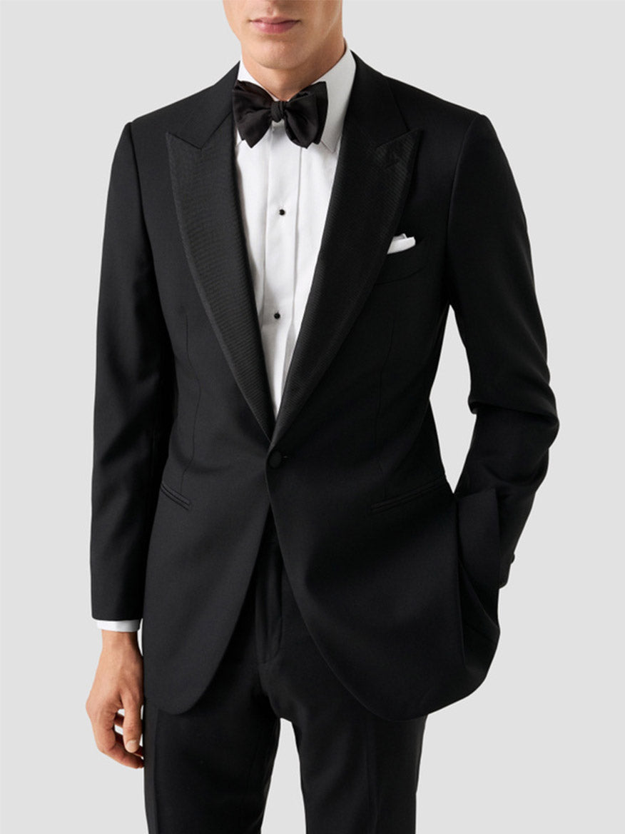 A man wears an Eton Contemporary Fit Fine Striped Bib Evening Shirt in White with French cuffs, styled with a black tuxedo, black bow tie, and pocket square. He stands against a plain background with one hand in his pocket.