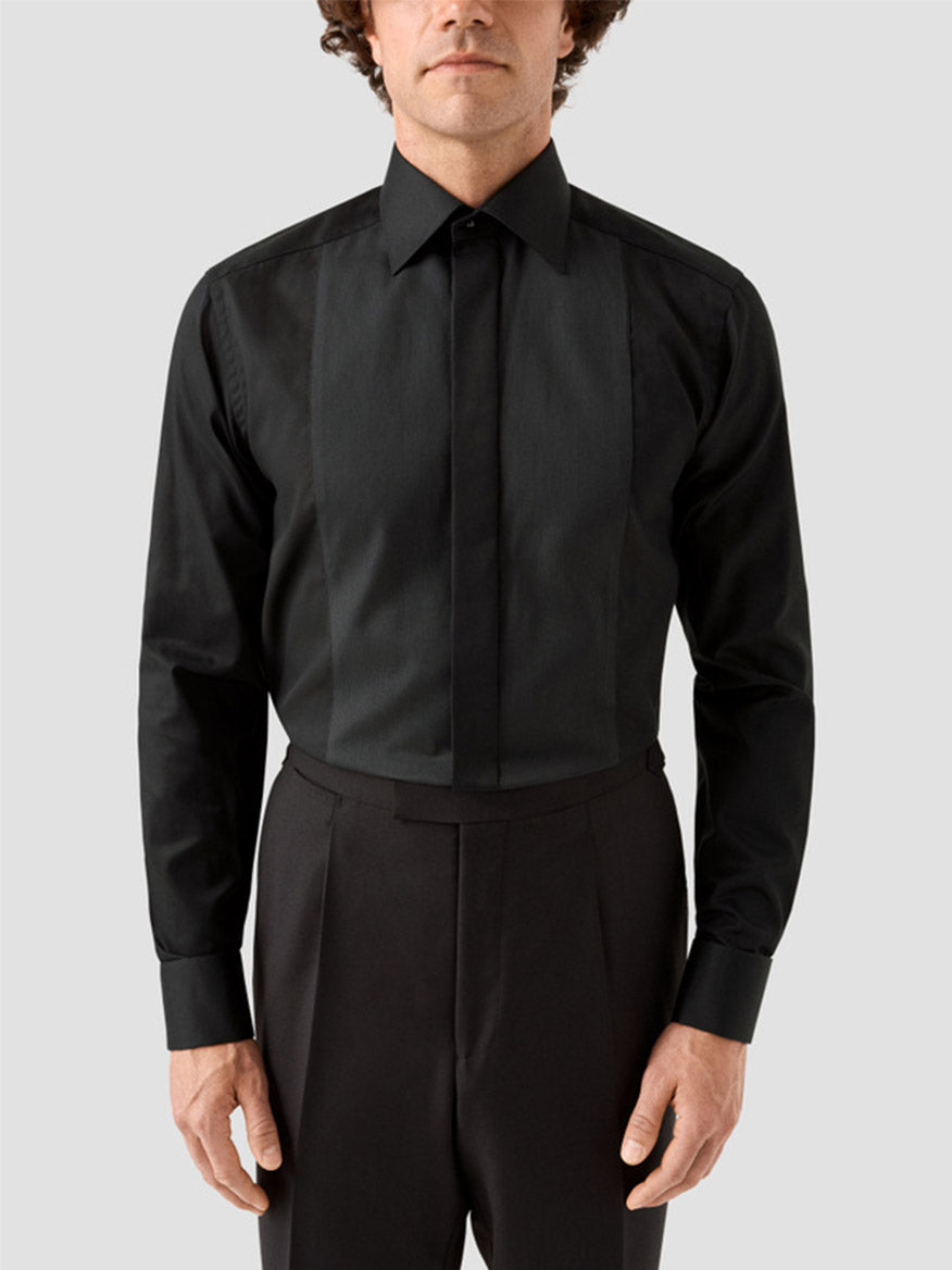 A man wearing the Eton Contemporary Fit Fine Striped Bib Evening Shirt in Black with French cuffs and black trousers stands against a plain light gray background.