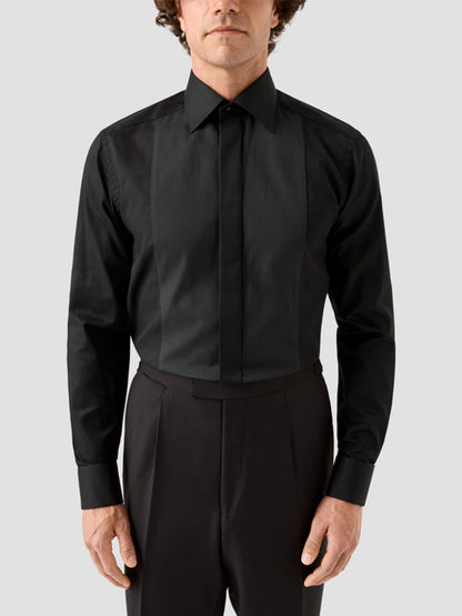 A man wearing the Eton Contemporary Fit Fine Striped Bib Evening Shirt in Black with French cuffs and black trousers stands against a plain light gray background.