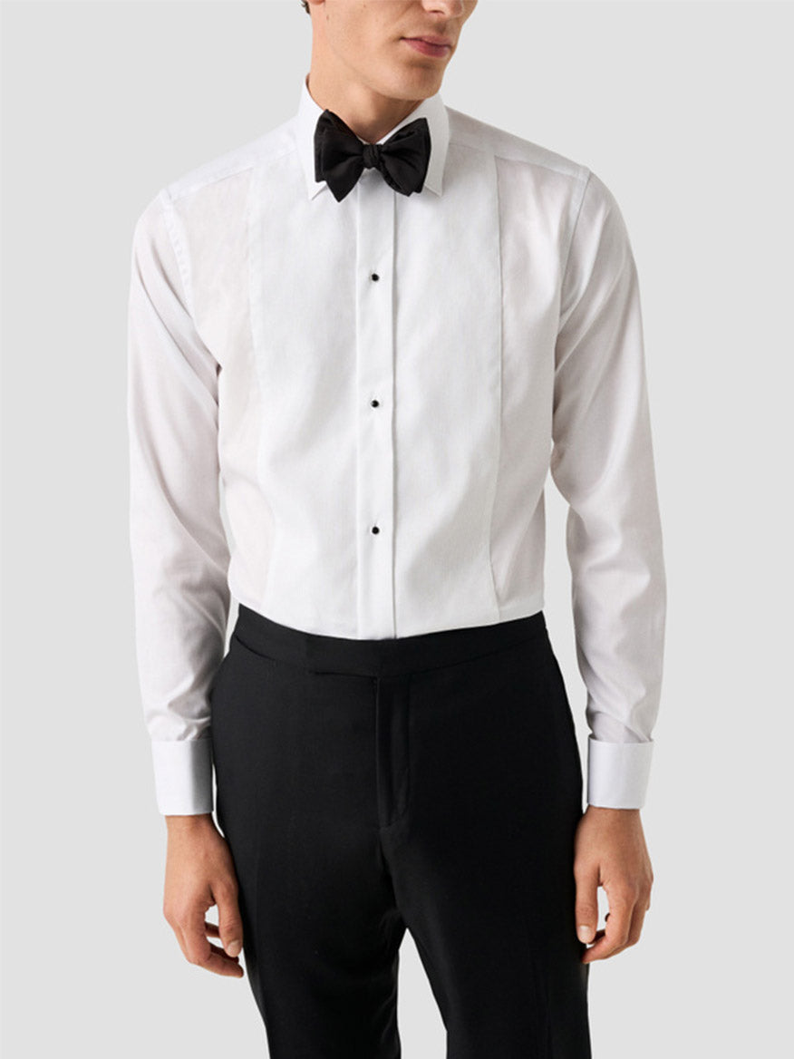 A man wearing the Eton Slim Fit Fine Striped Bib Evening Shirt in White with French cuffs, a black bow tie, and black trousers stands against a plain light gray background.