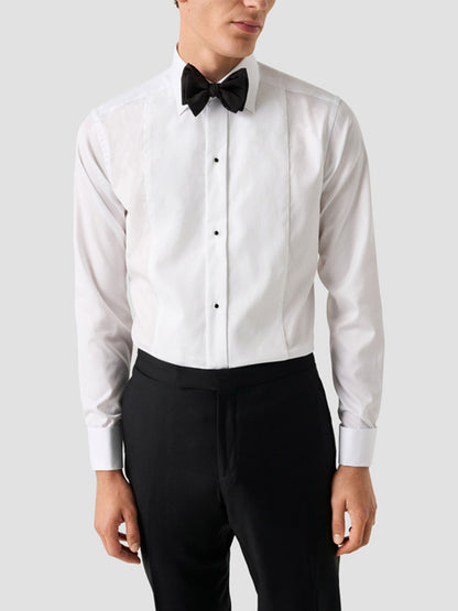 A man wearing the Eton Slim Fit Fine Striped Bib Evening Shirt in White with French cuffs, a black bow tie, and black trousers stands against a plain light gray background.