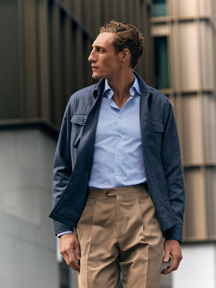 A man stands outdoors in an Eton Slim Fit Fine Striped Signature Twill Dress Shirt in Light Blue with a cutaway collar, paired with a dark blue jacket and beige pleated trousers—an elevated business classic look.