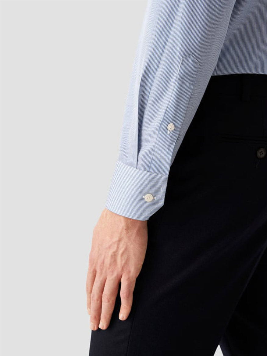 A person wearing the Eton Slim Fit Fine Striped Signature Twill Dress Shirt in Mid Blue and black pants, shown from the waist down with their arm relaxed by their side, embodying a business classic style.