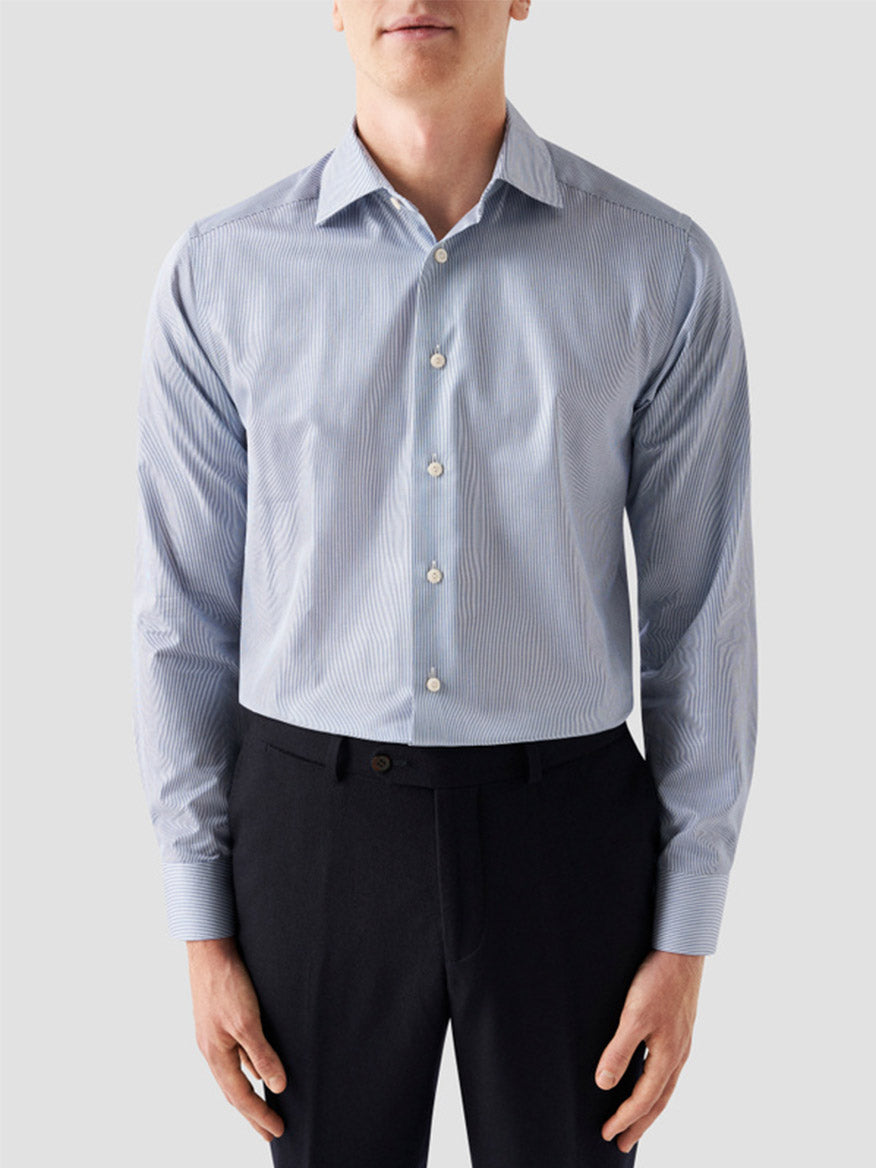 A man wearing the Eton Slim Fit Fine Striped Signature Twill Dress Shirt in Mid Blue with black dress pants stands against a plain light background, creating a classic business look.