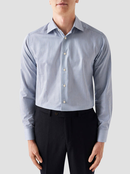 A man wearing the Eton Slim Fit Fine Striped Signature Twill Dress Shirt in Mid Blue with black dress pants stands against a plain light background, creating a classic business look.