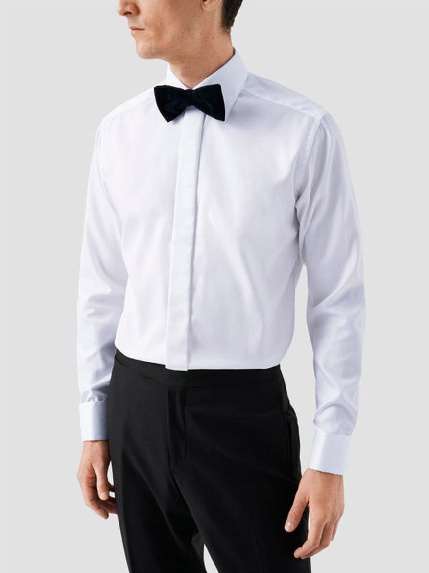 A man wearing an Eton Slim Fit Fly Front Evening Dobby Shirt in white, a black bow tie, and black trousers stands against a plain light grey background.