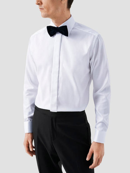 A man wearing an Eton Slim Fit Fly Front Evening Dobby Shirt in white, a black bow tie, and black trousers stands against a plain light grey background.