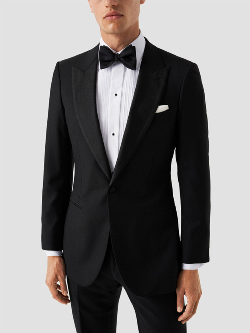A man wears the Eton Slim Fit Plissé Bib-Front Tuxedo Shirt in white, paired with a black tuxedo, black bow tie, and white pocket square, standing against a plain light gray background.