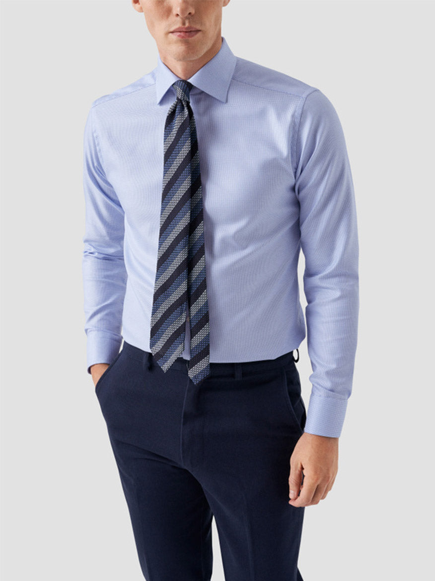 A man wearing the Eton Contemporary Fit Semi Solid Dobby Dress Shirt in Light Blue, a patterned tie, and dark navy trousers stands with one hand in his pocket against a plain background.