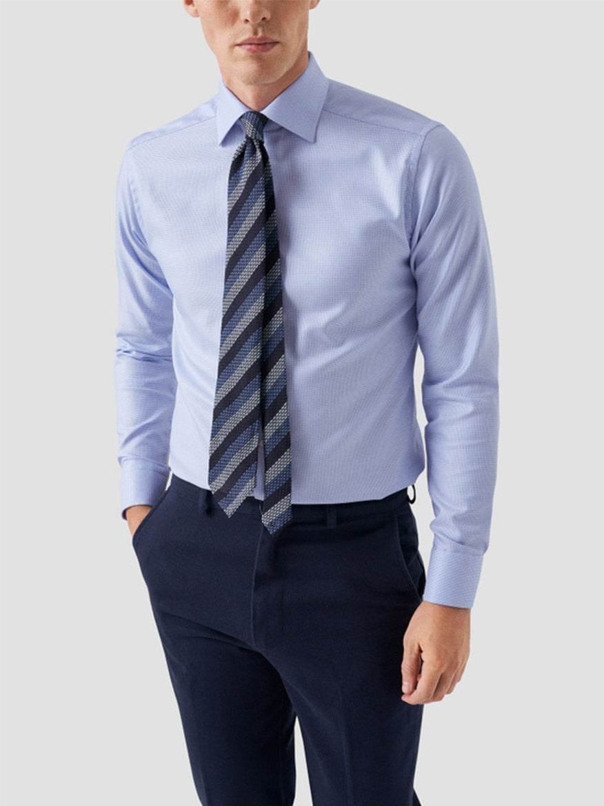 A man in an Eton Slim Fit Semi Solid Dobby Dress Shirt in light blue, navy dress pants, and a striped tie stands with one hand in his pocket against a plain light background.