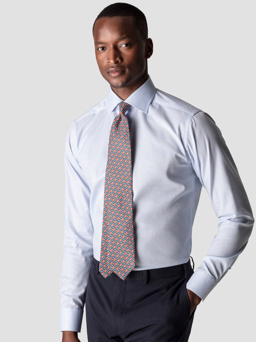 A man wears the Eton Slim Fit Semi Solid Piping Details Shirt in Light Blue, paired with a patterned tie and dark trousers, standing against a plain light gray background.