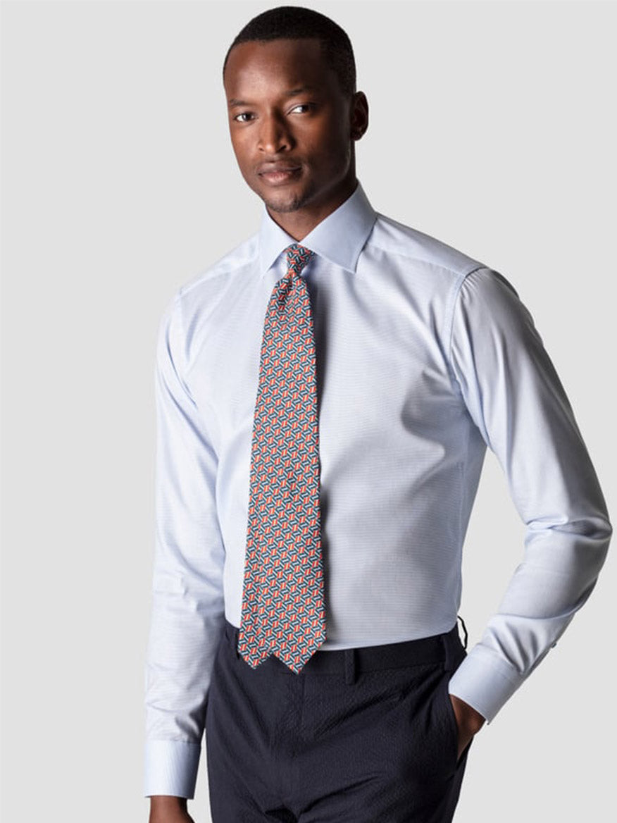 A man wears the Eton Contemporary Fit Semi Solid Piping Details Shirt in light blue, paired with a patterned tie and dark trousers, standing against a plain light gray background.