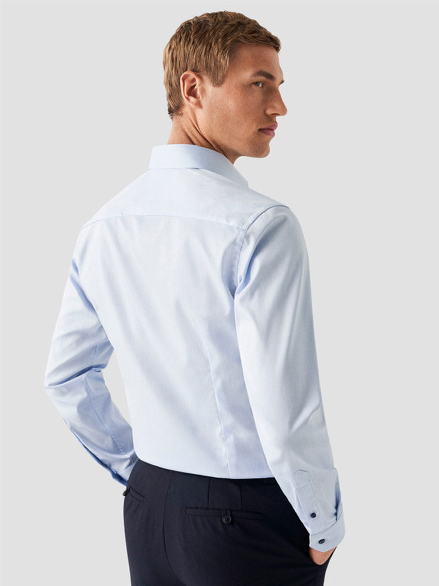 A man with short light brown hair, wearing an Eton Contemporary Fit Signature Twill Dress Shirt in light blue with navy details and dark trousers, stands facing away and glances over his left shoulder.
