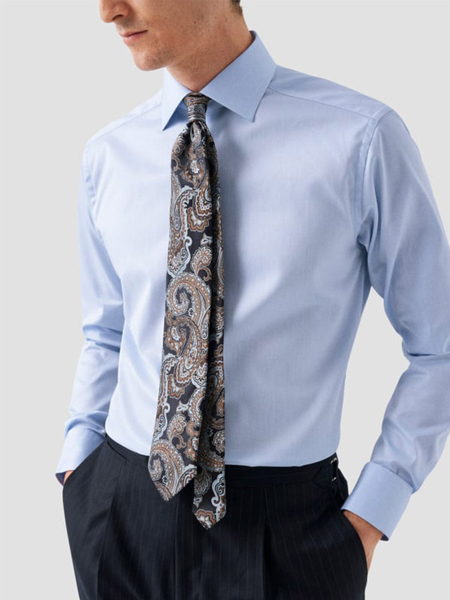 Man in an Eton Slim Fit Solid Signature Twill Dress Shirt in Light Blue - Single Cuff, paisley tie, and dark pinstripe trousers, standing with one hand in his pocket.