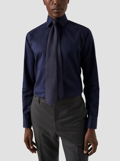 A person wearing an Eton Slim Fit Solid Signature Twill Dress Shirt in Navy Blue, paired with a matching tie and dark gray trousers, stands against a plain light gray background.