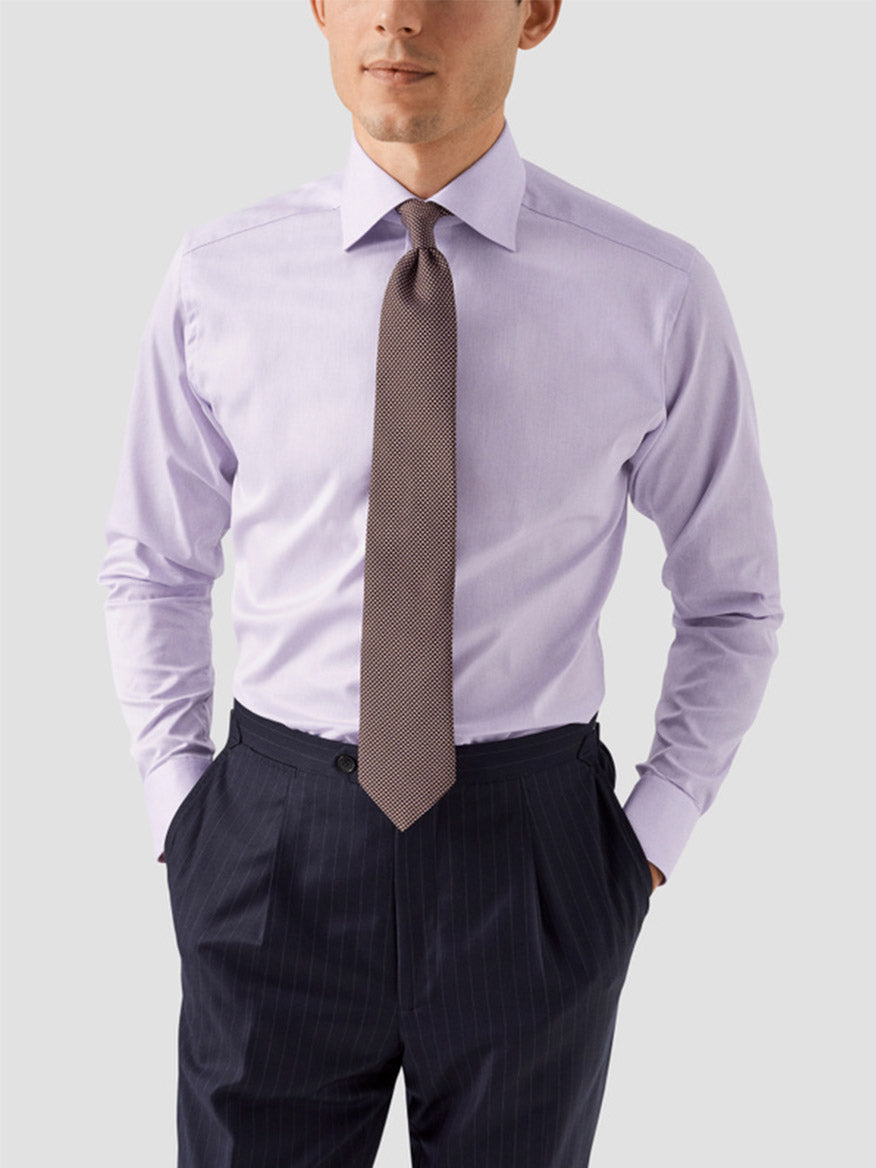 A man wears the Eton Slim Fit Solid Signature Twill Dress Shirt in Mid Purple, paired with a patterned tie and dark pinstripe trousers, standing hands-in-pockets and facing forward against a plain background.
