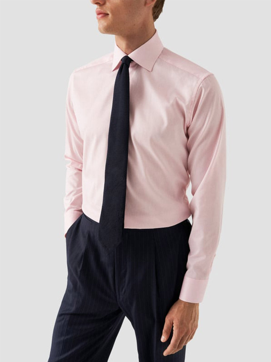 A man wears an Eton Slim Fit Solid Signature Twill Dress Shirt in Pink with a dark tie and pinstriped trousers, posing with one hand in his pocket against a plain background.