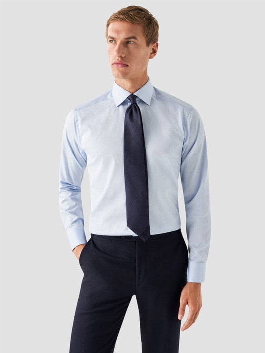 A man stands against a plain light gray background wearing an Eton Slim Fit Navy Details Signature Twill Dress Shirt in Light Blue, paired with a dark tie and matching dark trousers.