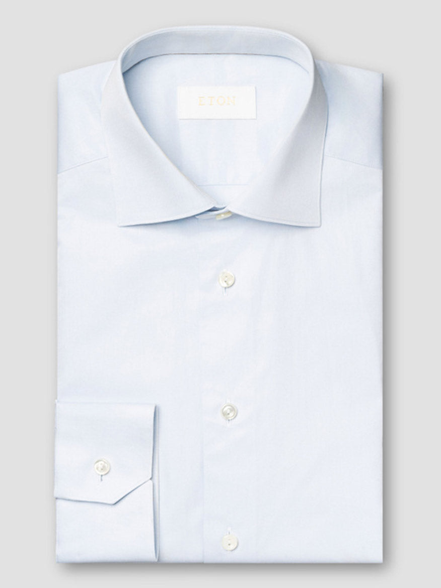 The Eton Slim Fit Solid Elevated Twill Dress Shirt in Light Blue, made from premium cotton, features white buttons and a visible brand label on the inside collar, displayed neatly folded against a light grey background.