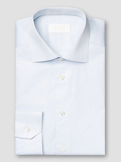 The Eton Slim Fit Solid Elevated Twill Dress Shirt in Light Blue, made from premium cotton, features white buttons and a visible brand label on the inside collar, displayed neatly folded against a light grey background.