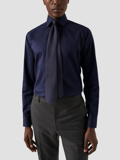 A person wearing an Eton Contemporary Fit Solid Signature Twill Dress Shirt in Navy Blue, paired with a matching tie and gray dress pants, stands against a plain light background.