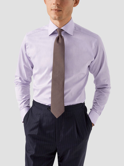 A man wears an Eton Contemporary Fit Solid Signature Twill Dress Shirt in Mid Purple, a patterned tie, and dark pinstripe trousers with his hands in his pockets against a plain light background.