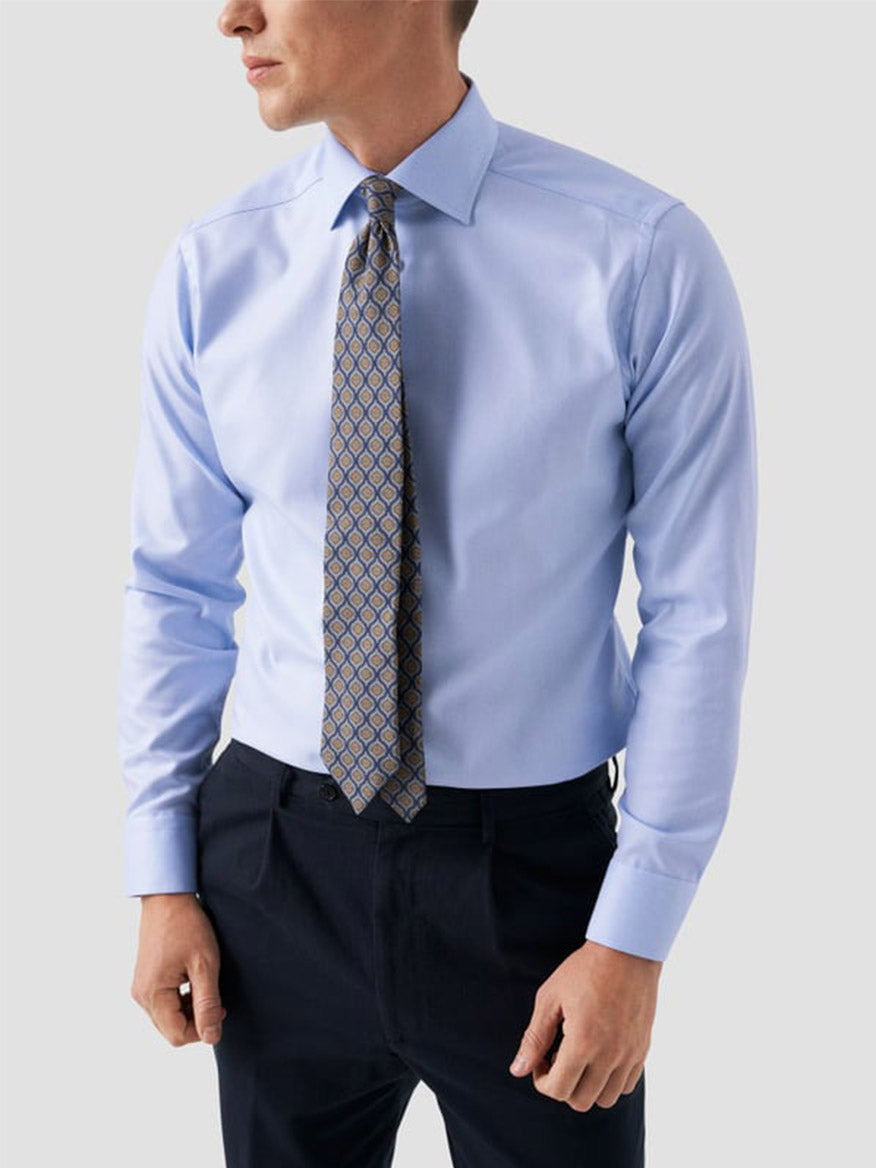 A man wears the Eton Contemporary Fit Solid Textured Twill Dress Shirt in Light Blue, paired with a patterned tie and dark dress pants, standing against a plain light background.