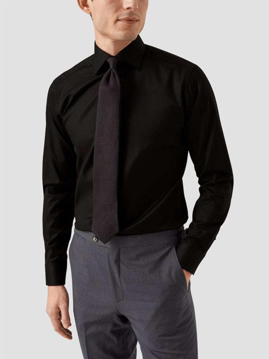 A man wears the Eton Contemporary Fit Solid Textured Twill Dress Shirt in Black, paired with a dark tie and gray dress pants. He stands with one hand in his pocket against a light background, exemplifying refined business style.