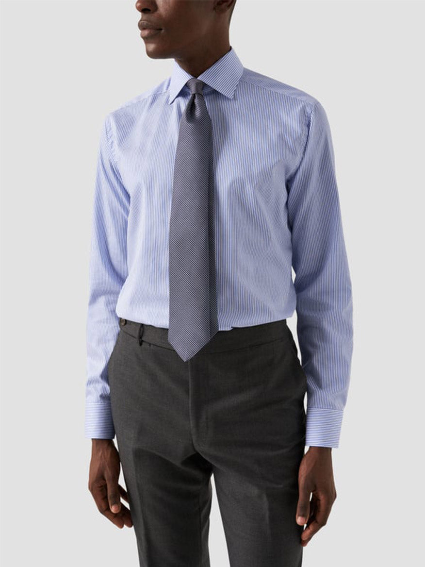 A man wearing an Eton Slim Fit Striped Fine Twill Dress Shirt in Mid Blue with a cut away collar, paired with dark gray trousers and a textured gray tie, stands against a plain light background.