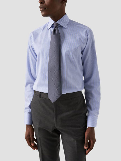 A man wearing an Eton Slim Fit Striped Fine Twill Dress Shirt in Mid Blue with a cut away collar, paired with dark gray trousers and a textured gray tie, stands against a plain light background.