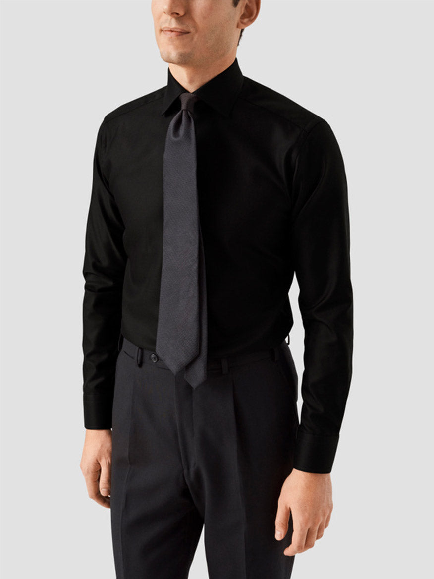 A man wears the Eton Slim Fit Black Textured Twill Dress Shirt with dark trousers and a textured gray tie, standing against a light gray background to showcase modern business style.