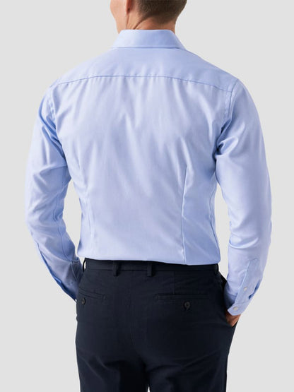 A man is seen from behind wearing an Eton Slim Fit Solid Textured Twill Dress Shirt in Light Blue with dark dress pants against a plain light gray background.