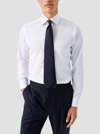 A man wears the Eton Slim Fit Signature Twill Dress Shirt in White with Grey Details, paired with dark striped dress pants and a dark tie. The wrinkle-resistant fabric keeps his look sharp all day as he stands against a plain background.